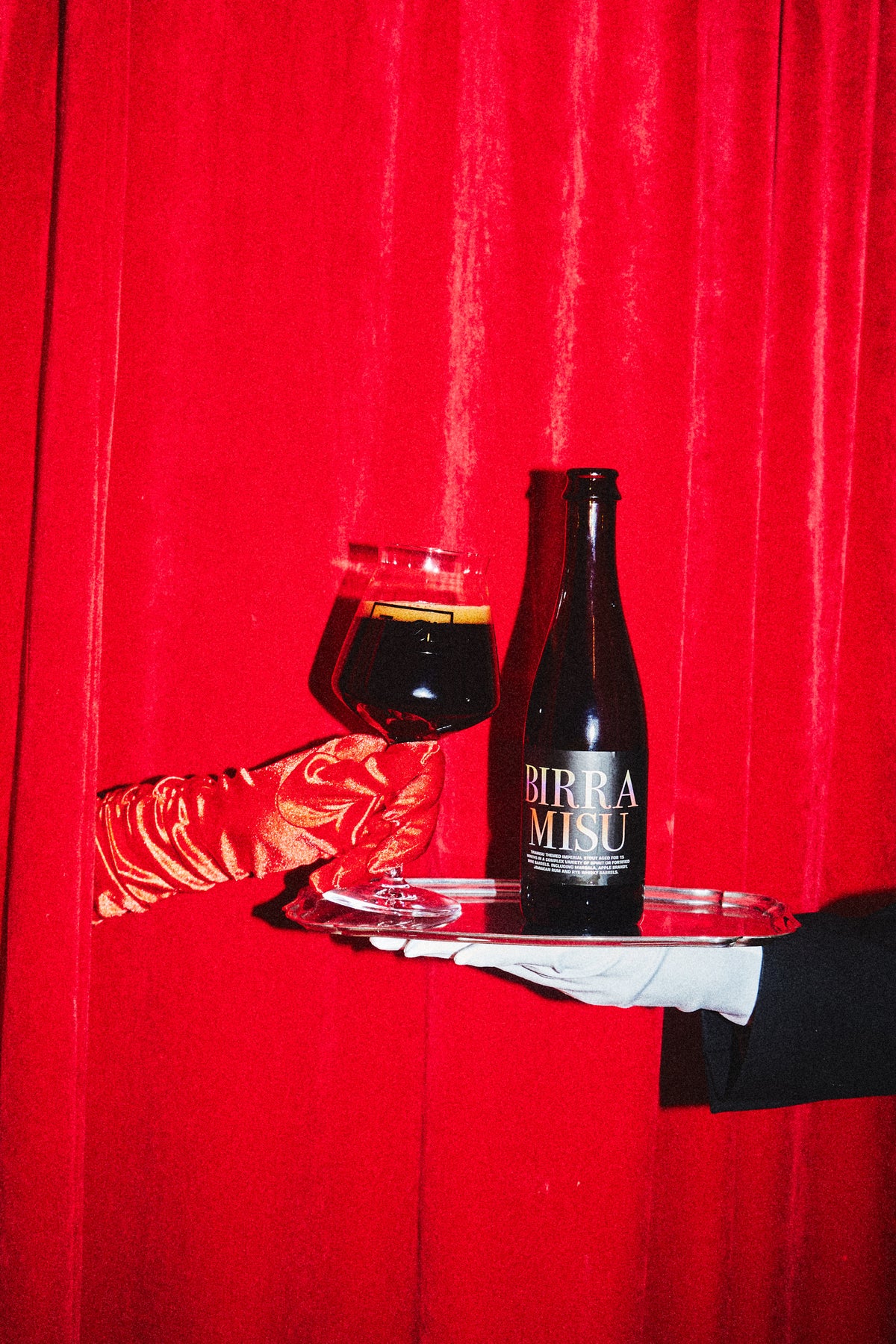 A bottle of Birramisu BA 2022 by To Øl and a filled beer glass on a silver tray, held by hands against a bright red curtain background.