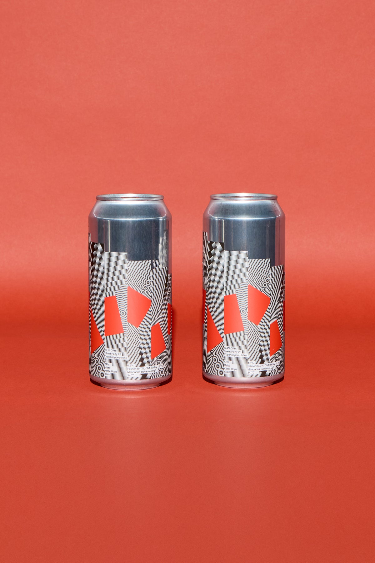 Two aluminum beverage cans with a black, white, and red geometric pattern, standing side-by-side on a solid red background.