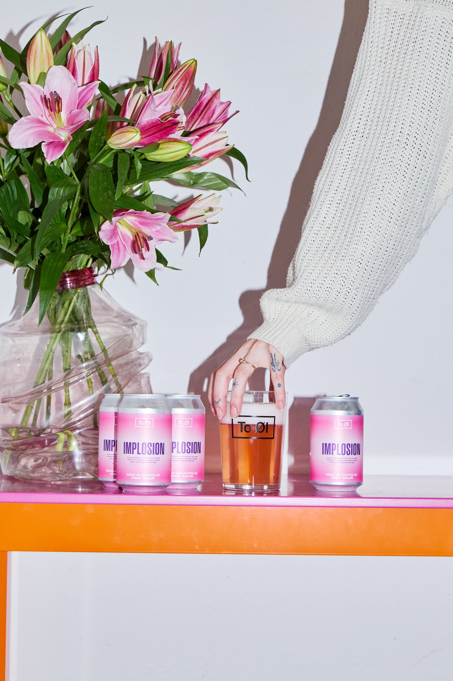 Three cans of To Øl's Implosion Pink Sour beer on a table with a glass and a bouquet of pink flowers.