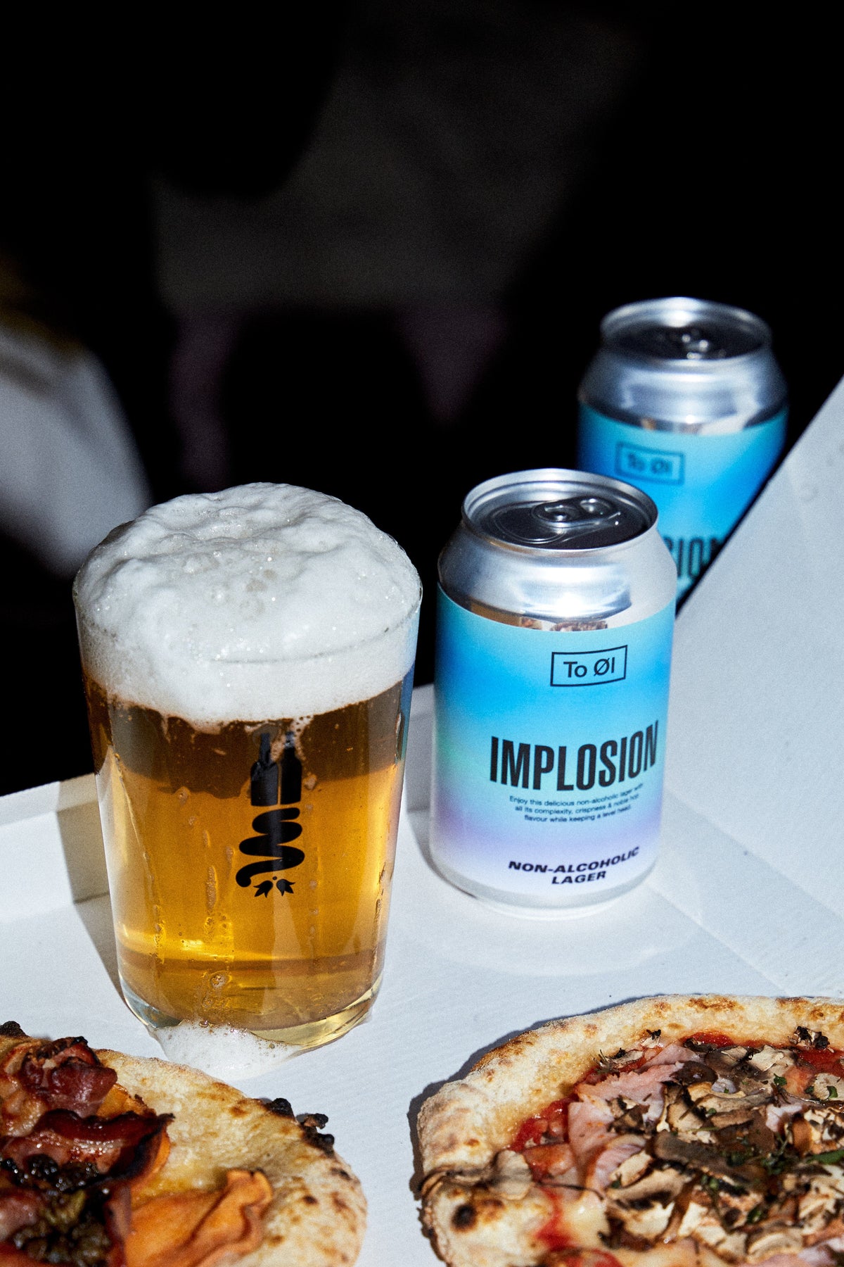 A glass of foamy golden beer and two To Øl Implosion Lager non-alcoholic canned beers on a white tray, with pizza slices partially visible in the foreground.