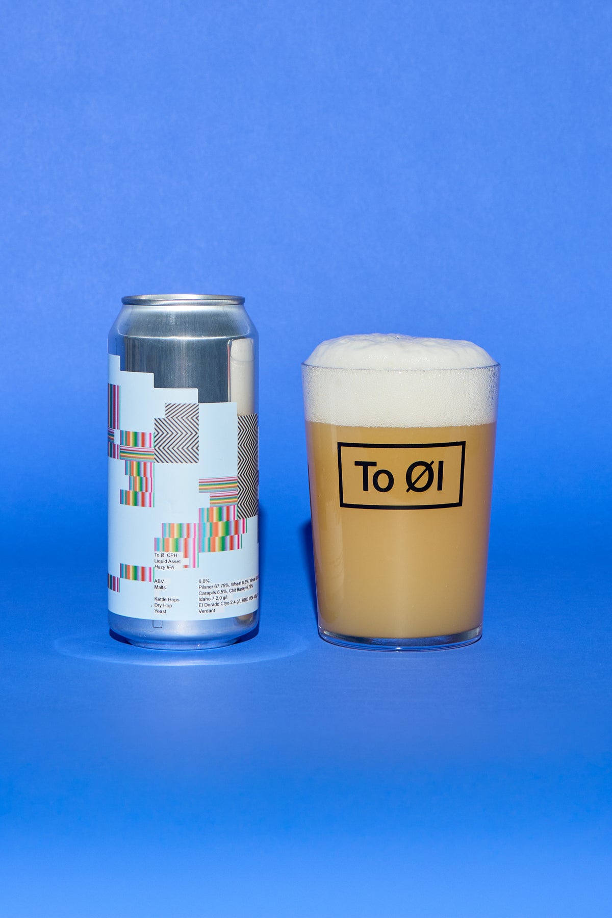 A silver aluminum can with a colorful geometric label stands next to a clear glass filled with a light tan beverage and topped with thick white foam. The glass has a black logo that reads 'To 01'.