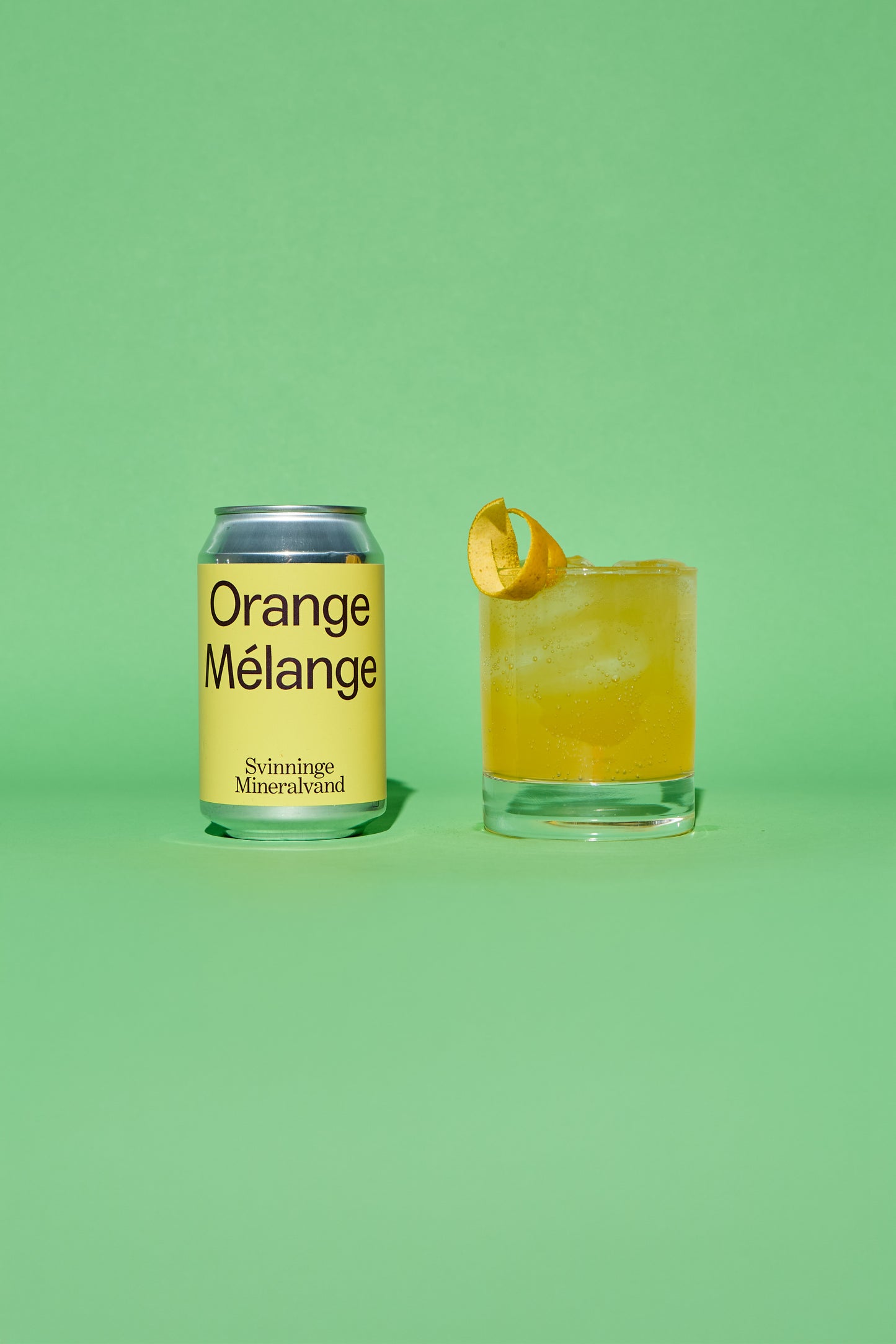 Yellow and silver can of 'Orange Melange' and a glass of orange cocktail on a green background.