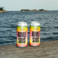 Two aluminum cans of To Øl Humle Sows Juicy IPA beer cans, featuring a yellow, pink, and orange gradient label, sitting on a weathered wooden dock with a body of water and distant land under a cloudy sky in the background.