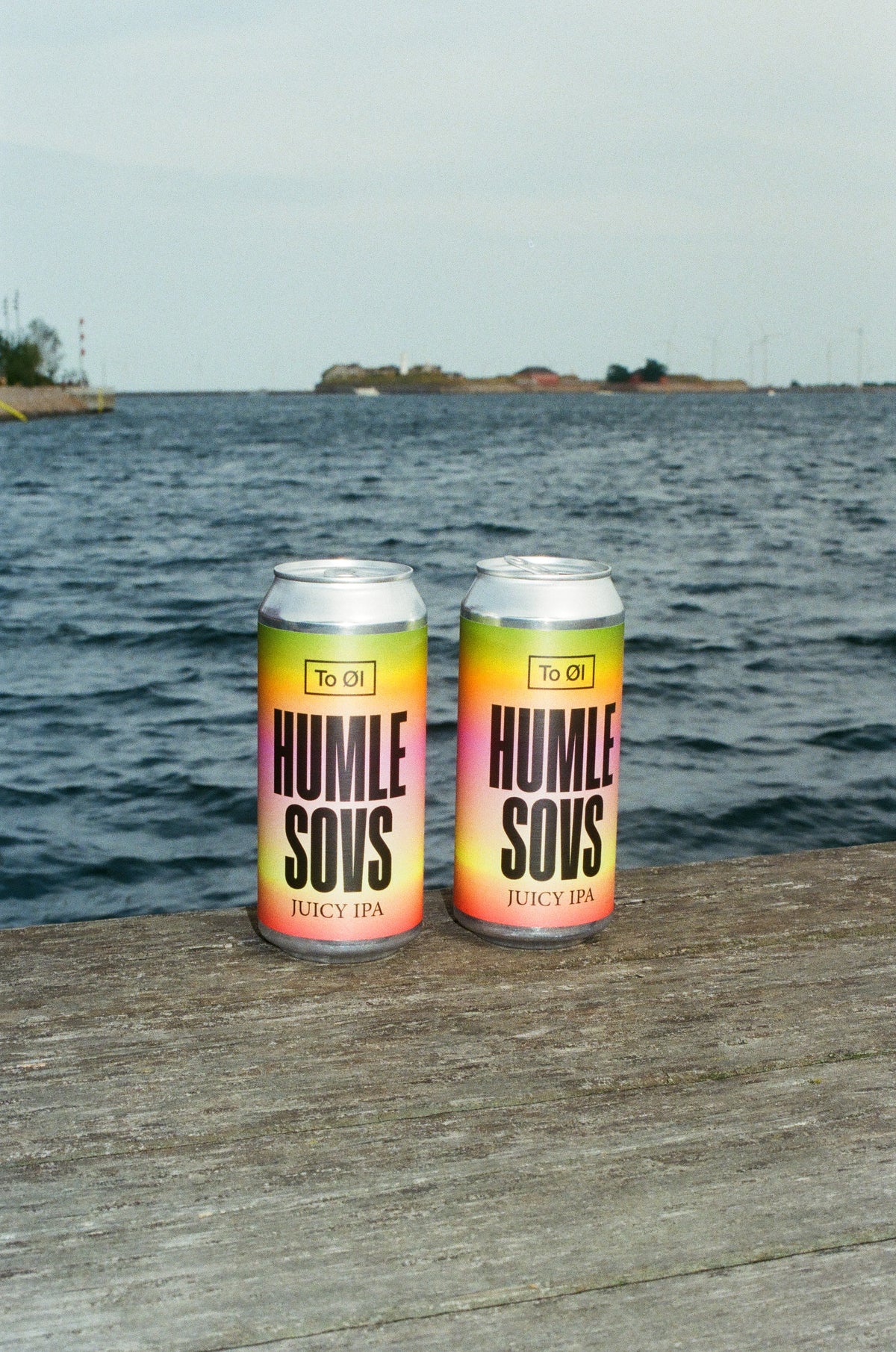 Two aluminum cans of To Øl Humle Sows Juicy IPA beer cans, featuring a yellow, pink, and orange gradient label, sitting on a weathered wooden dock with a body of water and distant land under a cloudy sky in the background.