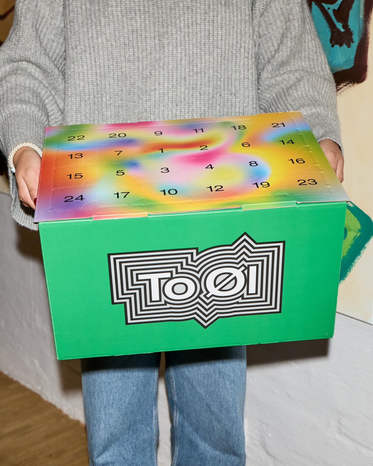 A person wearing a gray knit sweater and blue jeans holds a To Øl Advent Calendar - a green box with a colorful rainbow countdown calendar design on top and a large To Øl logo on the front.