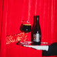 A bottle of Birramisu BA 2022 by To Øl and a filled beer glass on a silver tray, held by hands against a bright red curtain background.