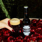 A hand with dark red nail polish holds a pint of dark stout beer with a thick head next to a dark bottle labeled 'To Øl Full Moon BA - 2024 ' on a wooden surface surrounded by shiny red ornaments, with a green velvet background.