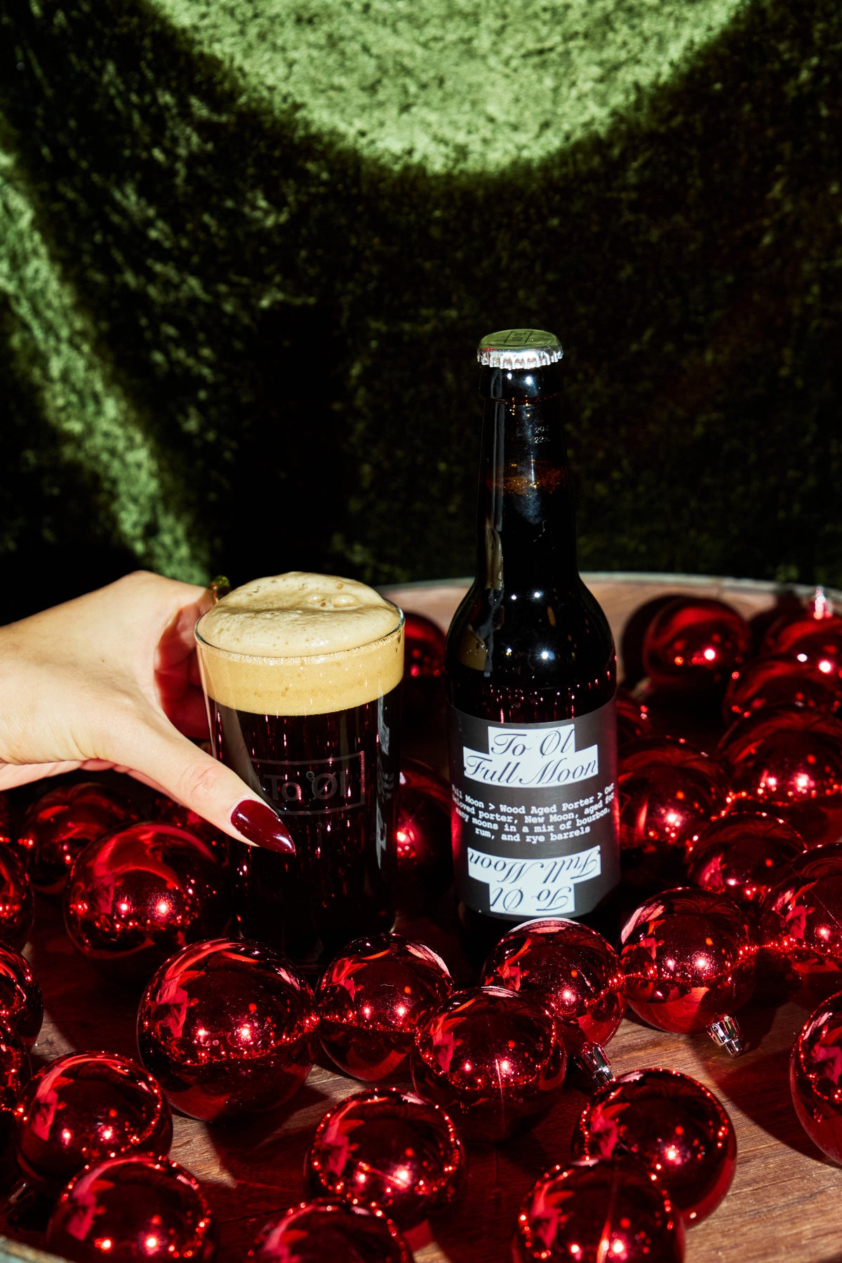 A hand with dark red nail polish holds a pint of dark stout beer with a thick head next to a dark bottle labeled 'To Øl Full Moon BA - 2024 ' on a wooden surface surrounded by shiny red ornaments, with a green velvet background.