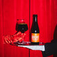 A bottle of To Øl Goliat dark beer with an orange label and a glass of the same dark beer, presented on a silver tray held by a hand in a white glove against a red curtain background.