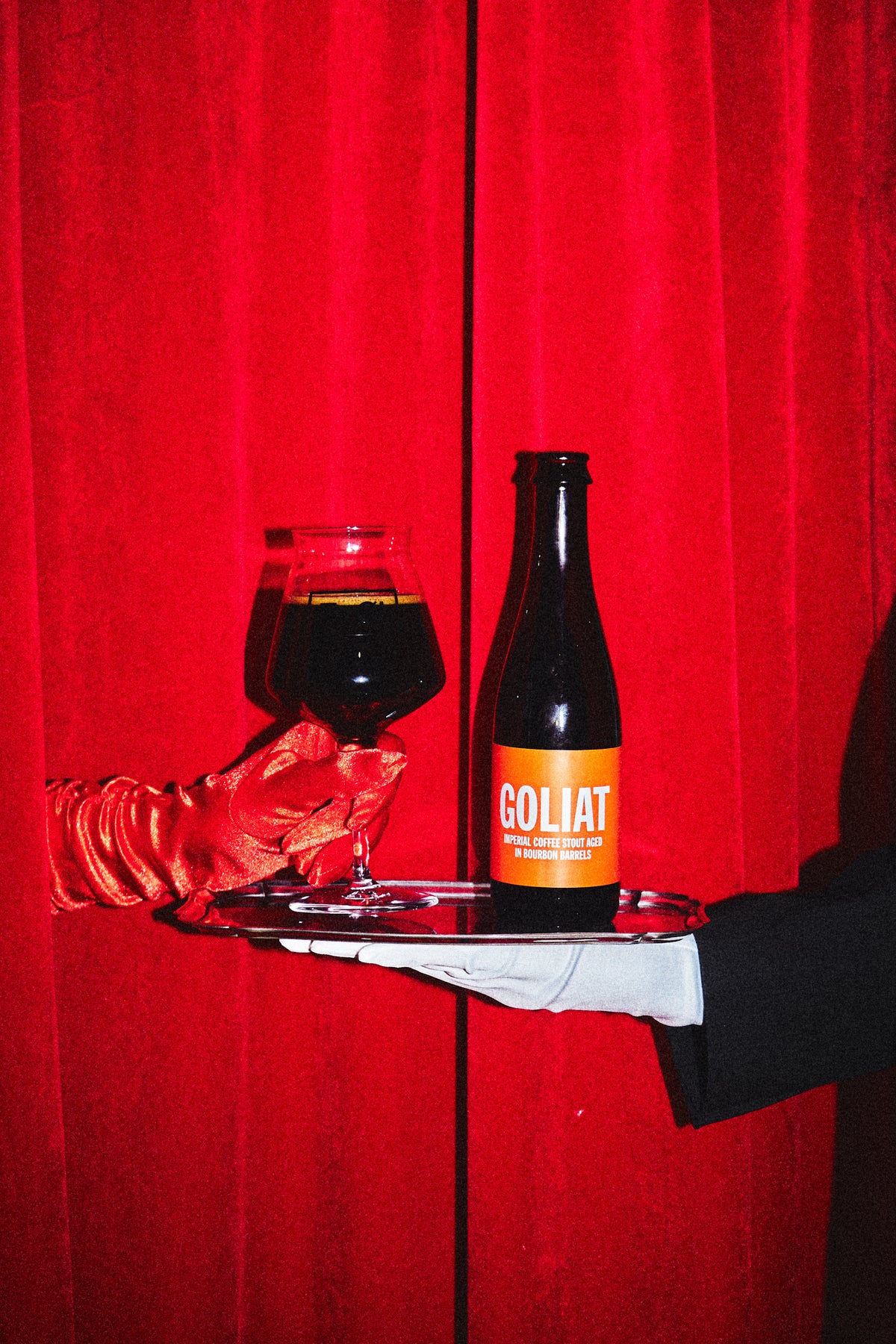 A bottle of To Øl Goliat dark beer with an orange label and a glass of the same dark beer, presented on a silver tray held by a hand in a white glove against a red curtain background.