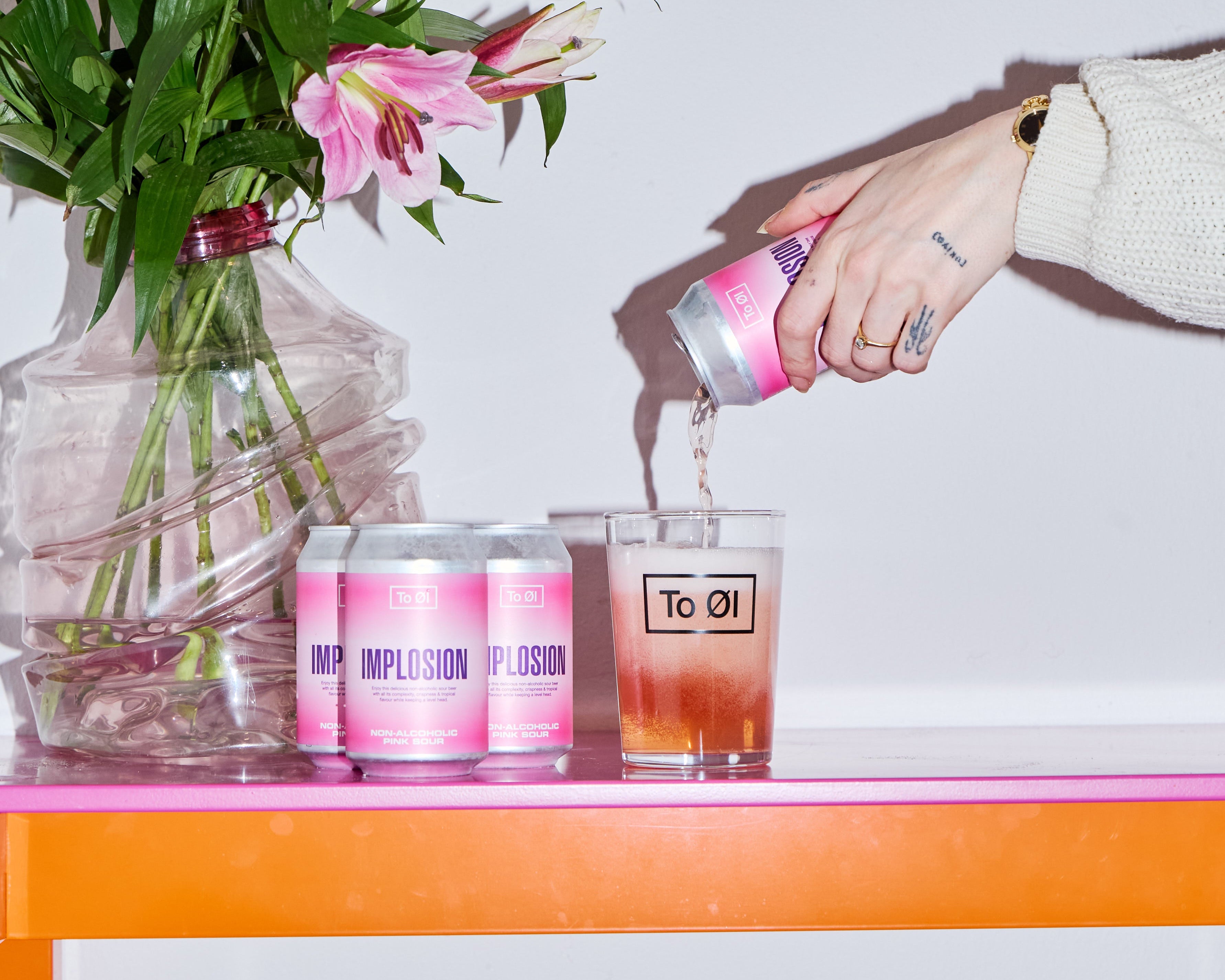 Person pouring a drink into a glass with flowers and bottles on an orange table.