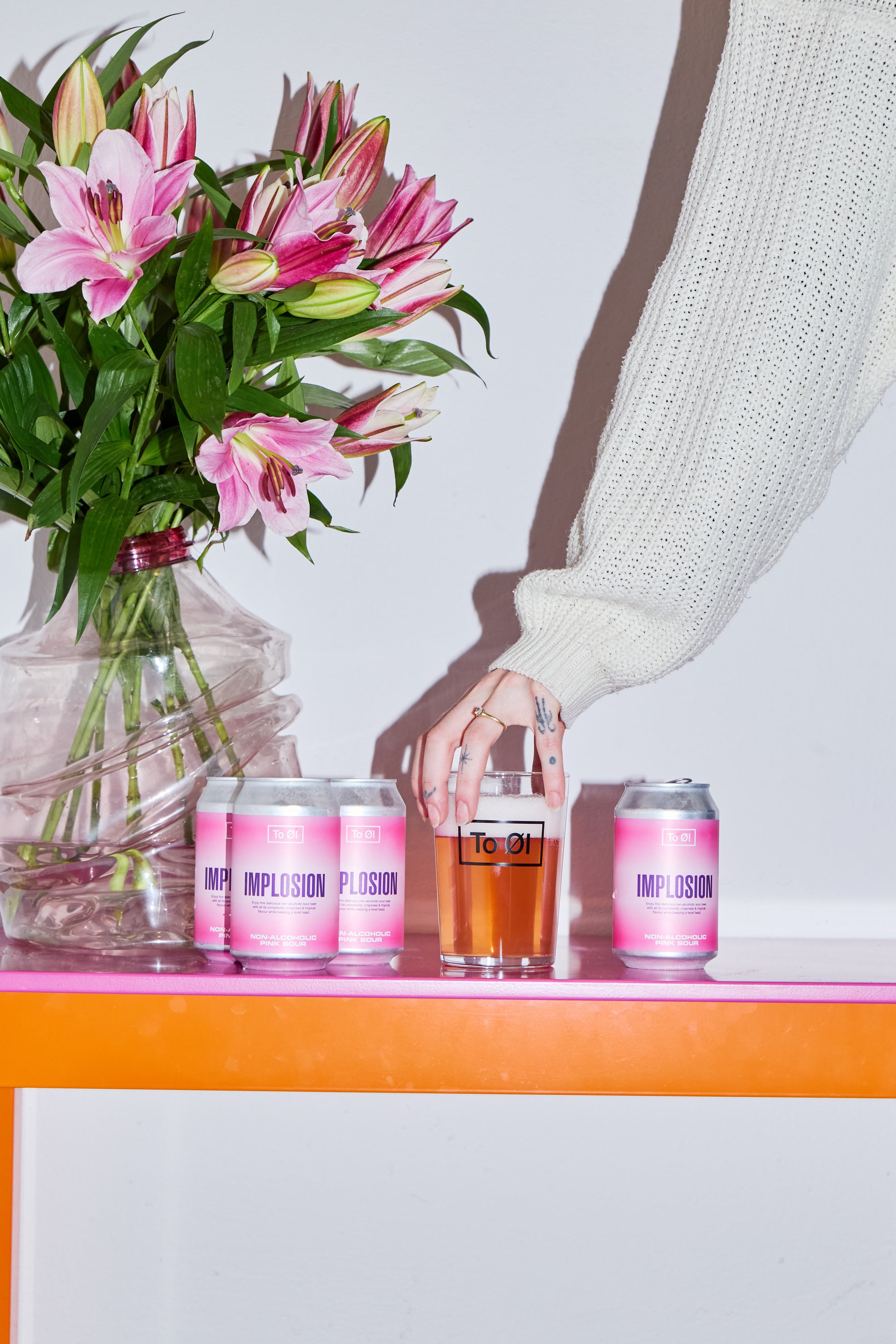 Three cans of To Øl's Implosion Pink Sour beer on a table with a glass and a bouquet of pink flowers.