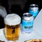 A glass of foamy golden beer and two To Øl Implosion Lager non-alcoholic canned beers on a white tray, with pizza slices partially visible in the foreground.