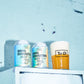 Two cans of Whirl Domination India Pale Ale lined up on a white ledge against a light blue brick wall, with a pint of amber beer in a glass labeled To Øl standing next to them.