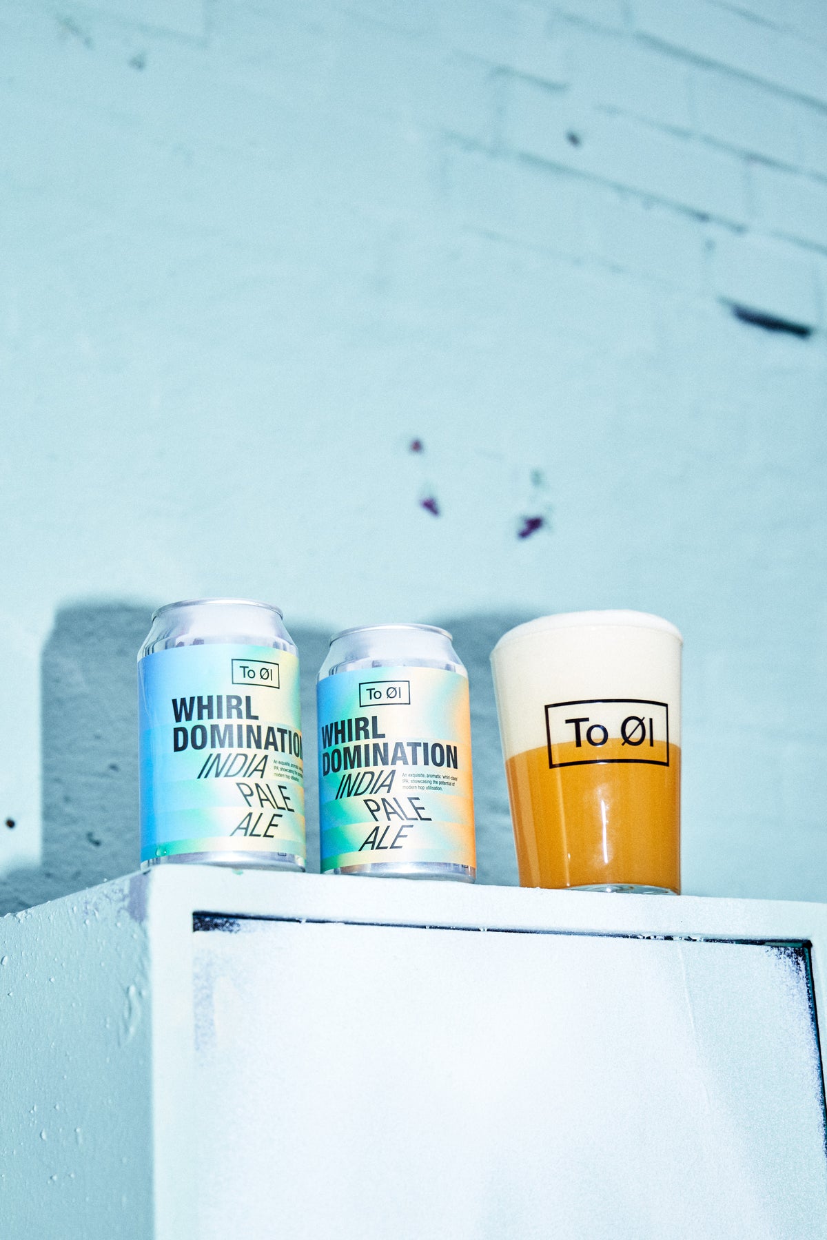Two cans of Whirl Domination India Pale Ale lined up on a white ledge against a light blue brick wall, with a pint of amber beer in a glass labeled To Øl standing next to them.