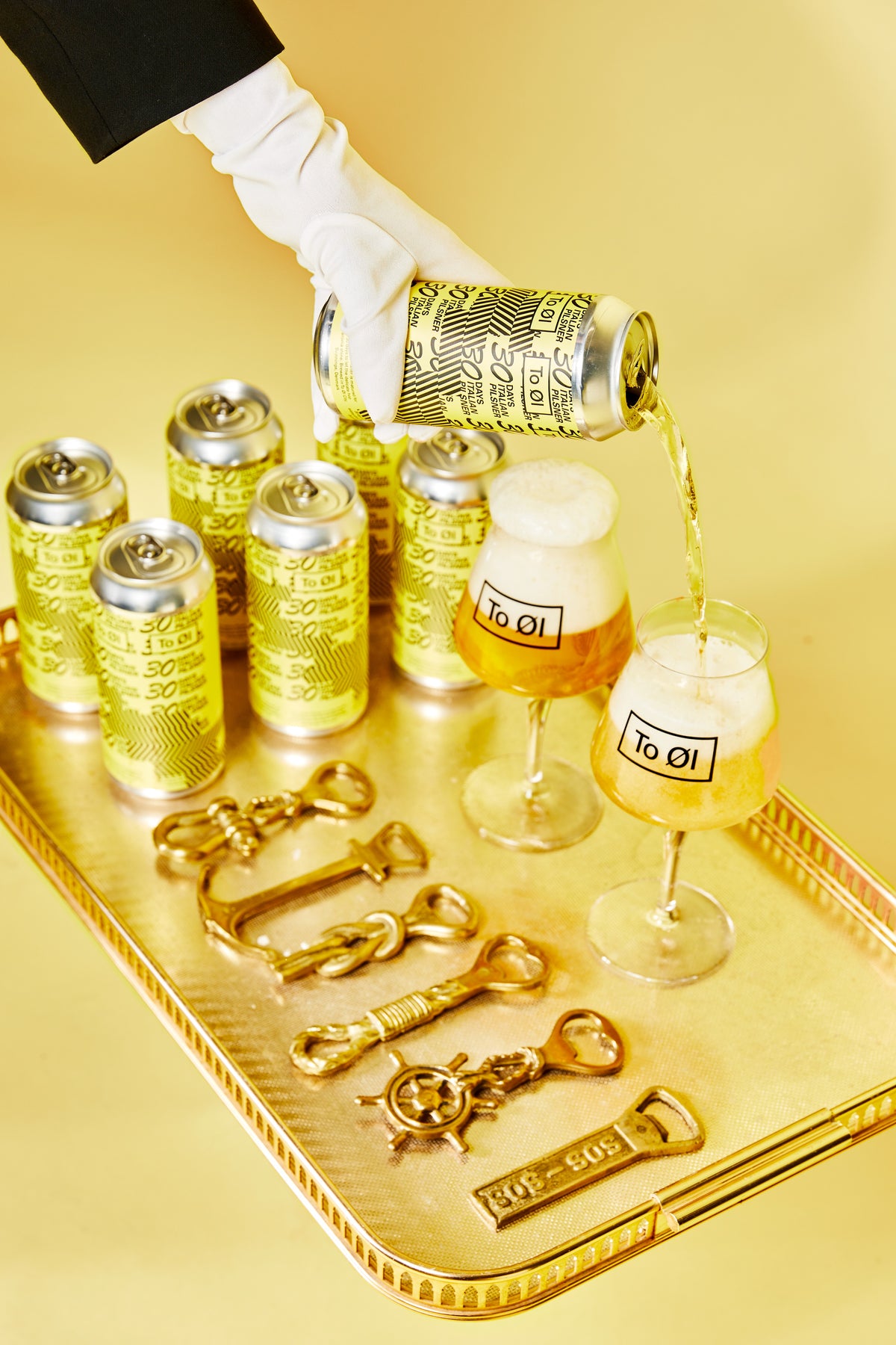 A hand in a white glove pours a golden beverage from a metallic gold can onto two glasses with To Øl Italian Pilsner labels, arranged on a gold tray with matching gold cans and decorative gold bottle opener keychains.