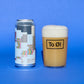 A silver aluminum can with a colorful geometric label stands next to a clear glass filled with a light tan beverage and topped with thick white foam. The glass has a black logo that reads 'To 01'.