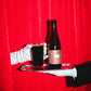 A dark brown glass bottle oand a matching dark drink in a clear glass, both held on a silver tray by gloved hands against a bright red curtain background. The bottle label reads 'Mexican Hot Chocolat BA Imperial Stout 2022' by To Øl.