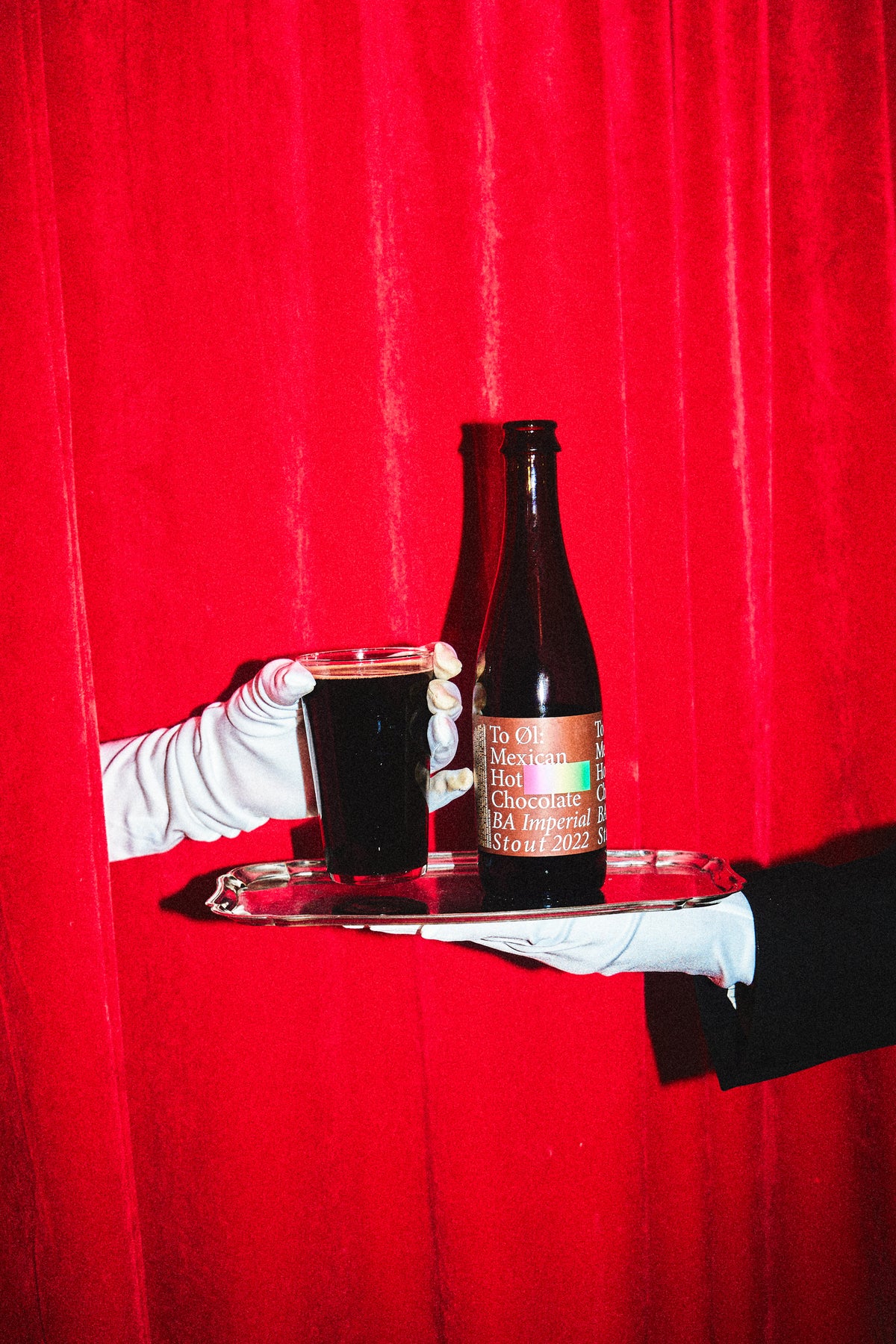 A dark brown glass bottle oand a matching dark drink in a clear glass, both held on a silver tray by gloved hands against a bright red curtain background. The bottle label reads 'Mexican Hot Chocolat BA Imperial Stout 2022' by To Øl.