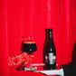A dark bottle of Mexican Hot Chocolat BA Imperial Stout 2022 and a filled glass on a silver tray, held by a hand in a red satin glove and another in a white sleeve, set against a bright red velvet curtain.