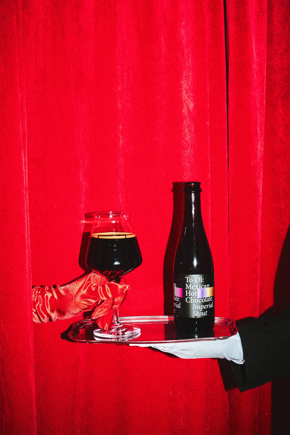 A dark bottle of Mexican Hot Chocolat BA Imperial Stout 2022 and a filled glass on a silver tray, held by a hand in a red satin glove and another in a white sleeve, set against a bright red velvet curtain.