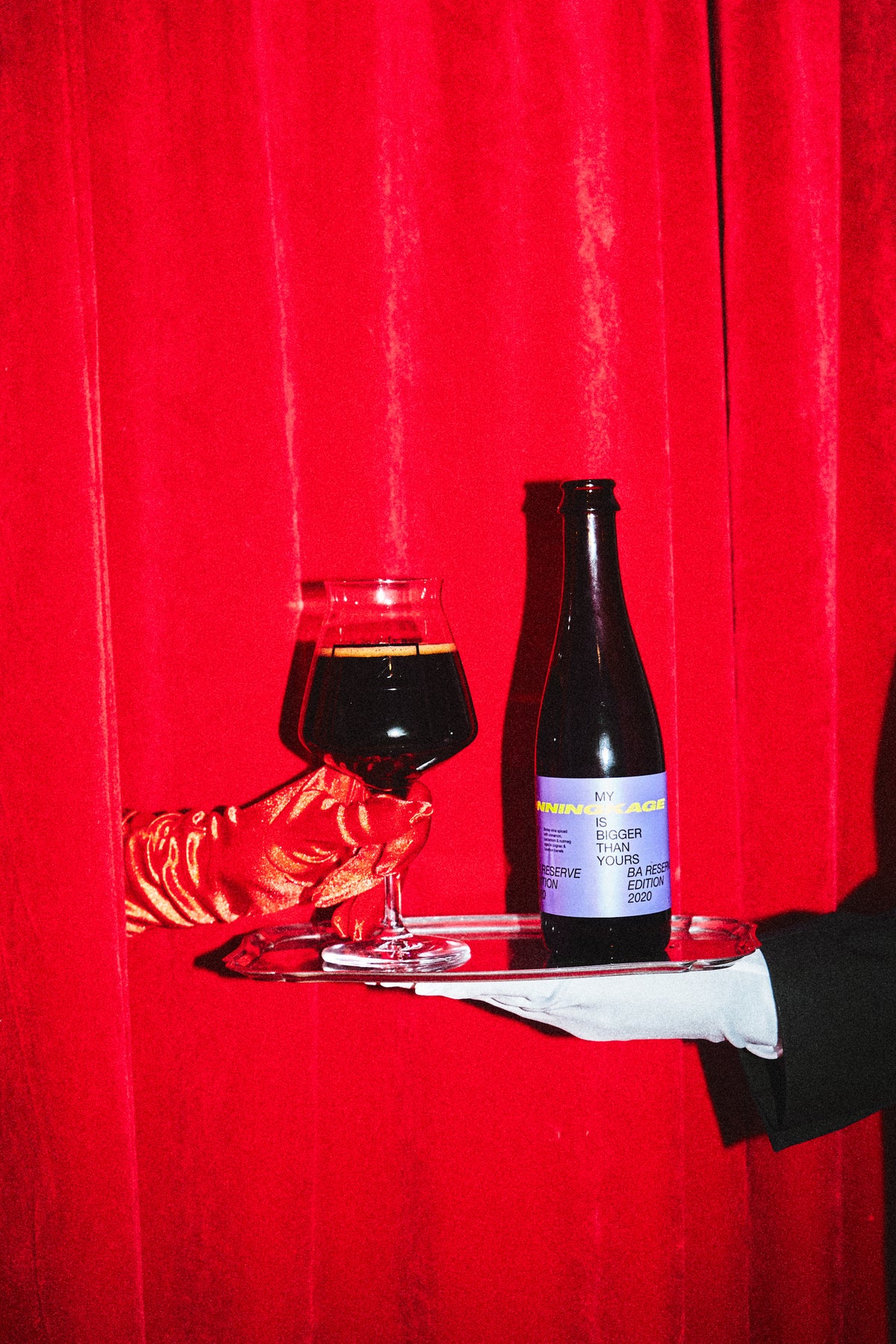 A bottle of To Øl My Honningskage, and a filled glass on a silver tray, held by a hand in a white glove against a bright red curtain background.