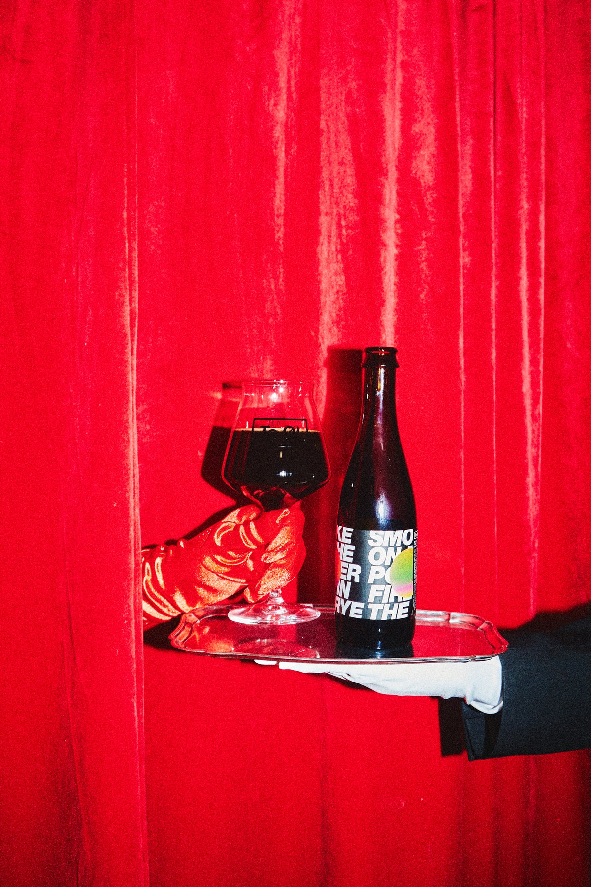 One bottle of Smoke On The Porter, Fire In The Rye - BA 2022 on a silver tray with a red glove holding a beer glass filled of the dark beer on a background with red curtains.