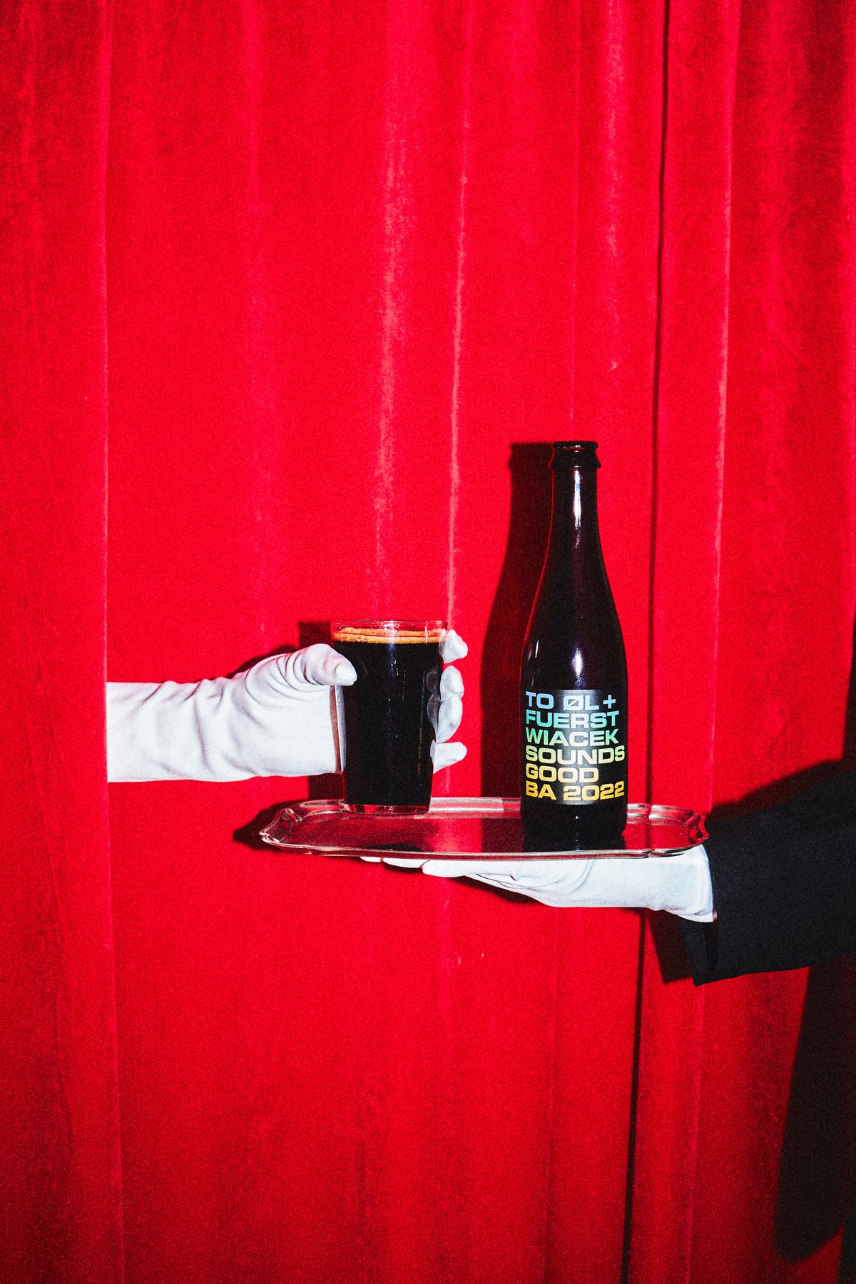 A dark beer bottle - Sounds Good BA 2022 by To Øl x Fuerst Wiacek, and matching dark pint glass on a silver tray, held by gloved hands against a bright red curtain backdrop. The bottle has colorful text label with color gradient.