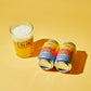 Two cans of To Øl Snuble Juice and a glass of the golden beverage with foam, arranged on a solid yellow background.