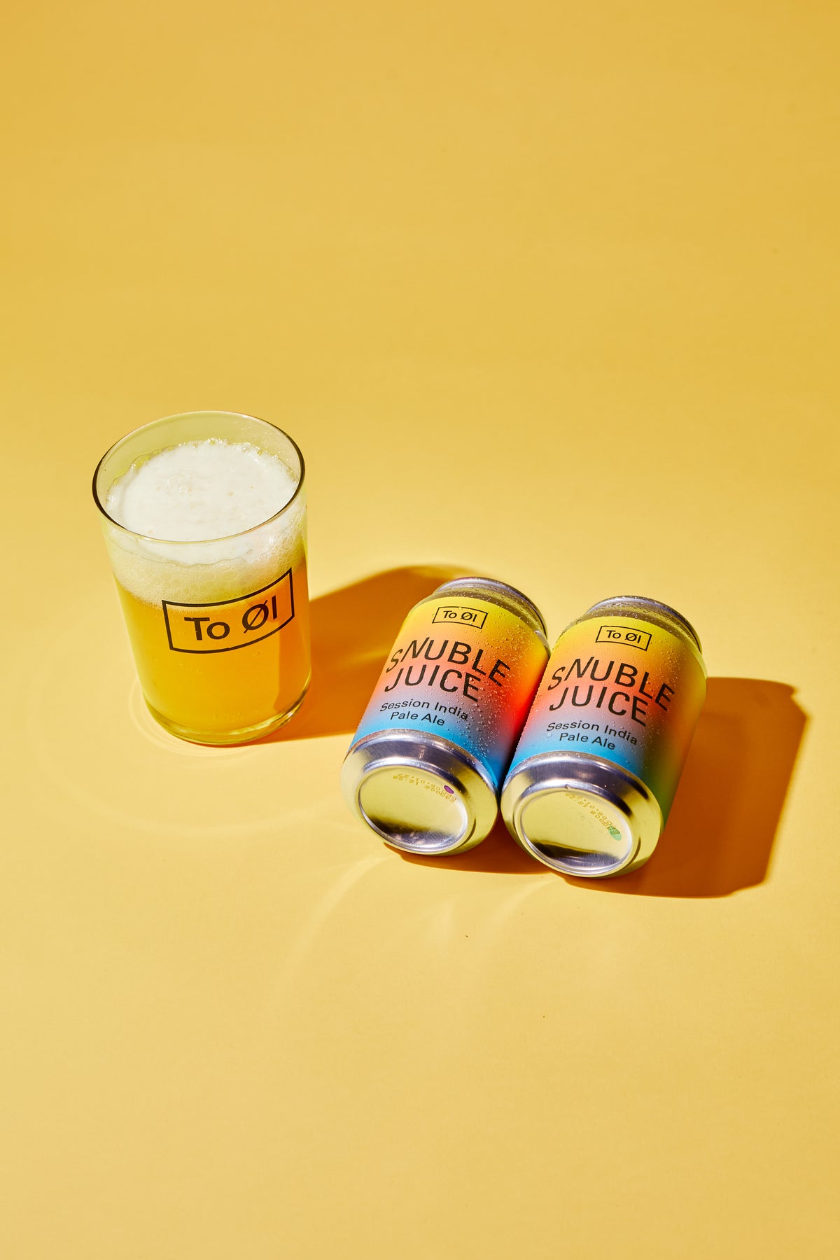 Two cans of To Øl Snuble Juice and a glass of the golden beverage with foam, arranged on a solid yellow background.