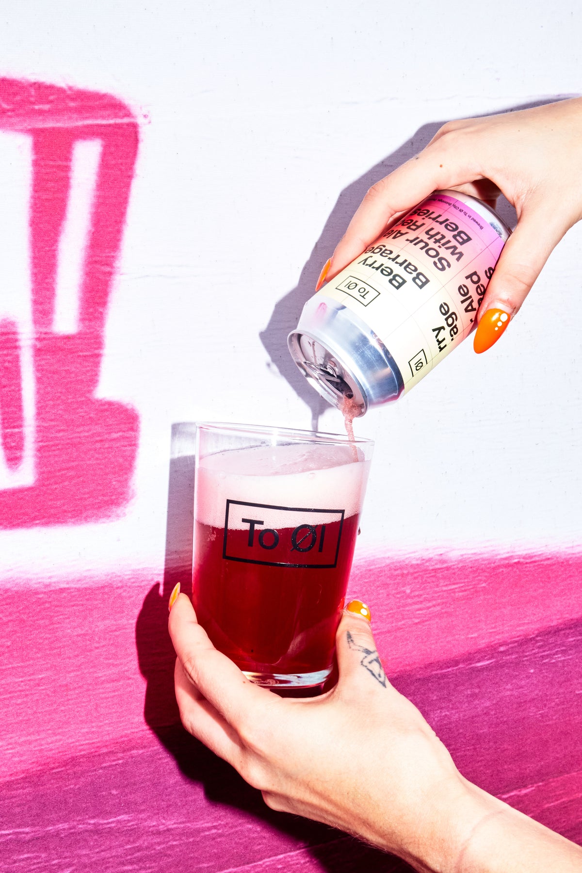 Person pouring a drink from a To Øl Berry Barrage can into a glass with a pink background.