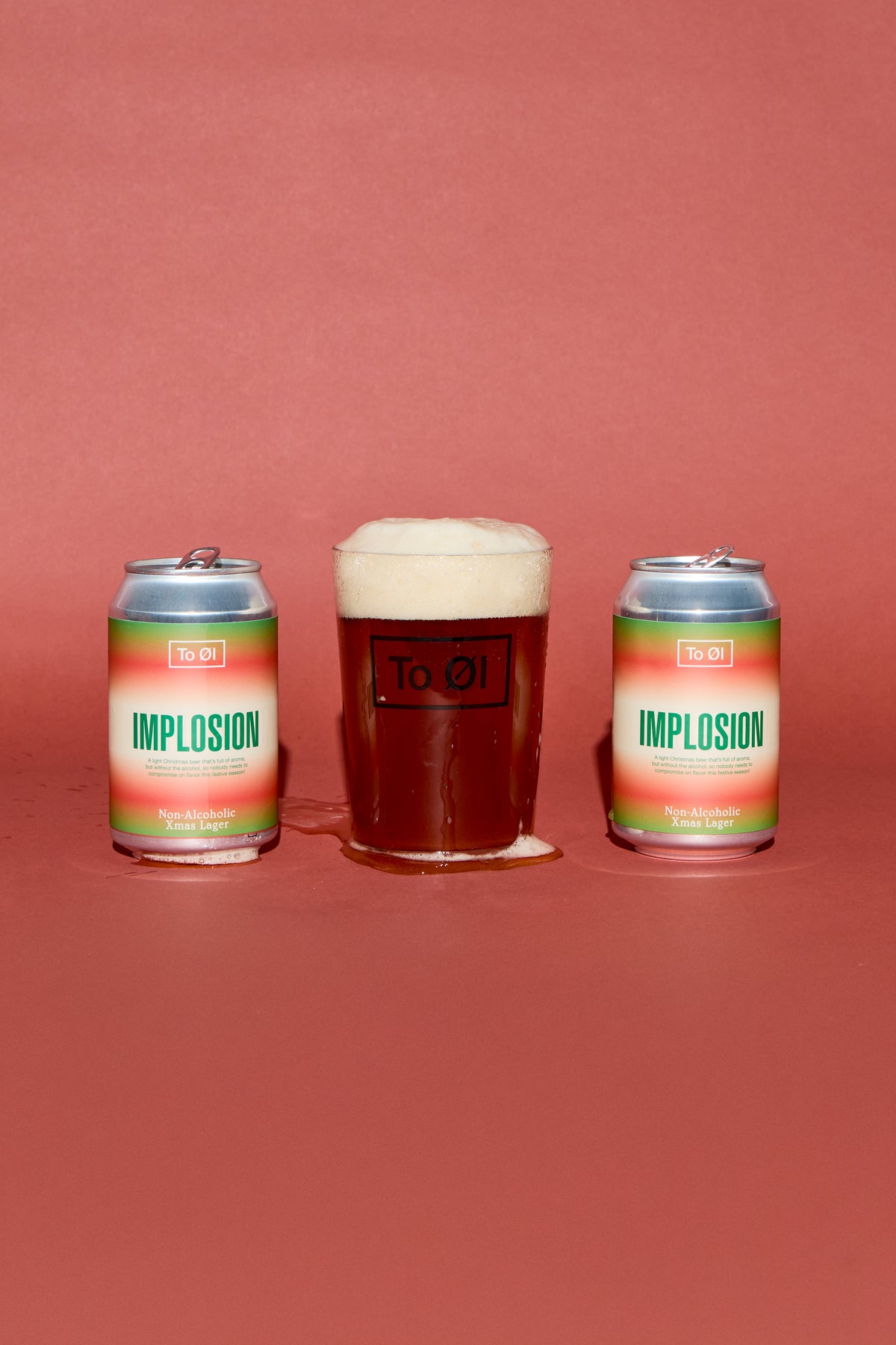 Two aluminum cans of To:01 Implosion beer flanked by a glass of dark amber beer with a thick white head, set against a solid terracotta background.