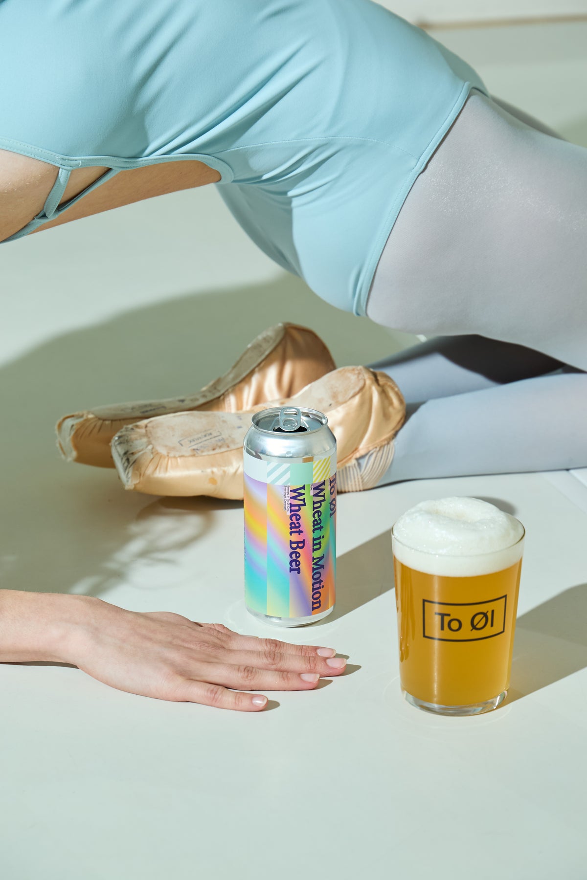 A lifestyle shot featuring a can of To Øl Wheat in Motion Beer IPA next to a glass of beer with foam, styled with ballet shoes and a dancer's hand and torso on a white surface.