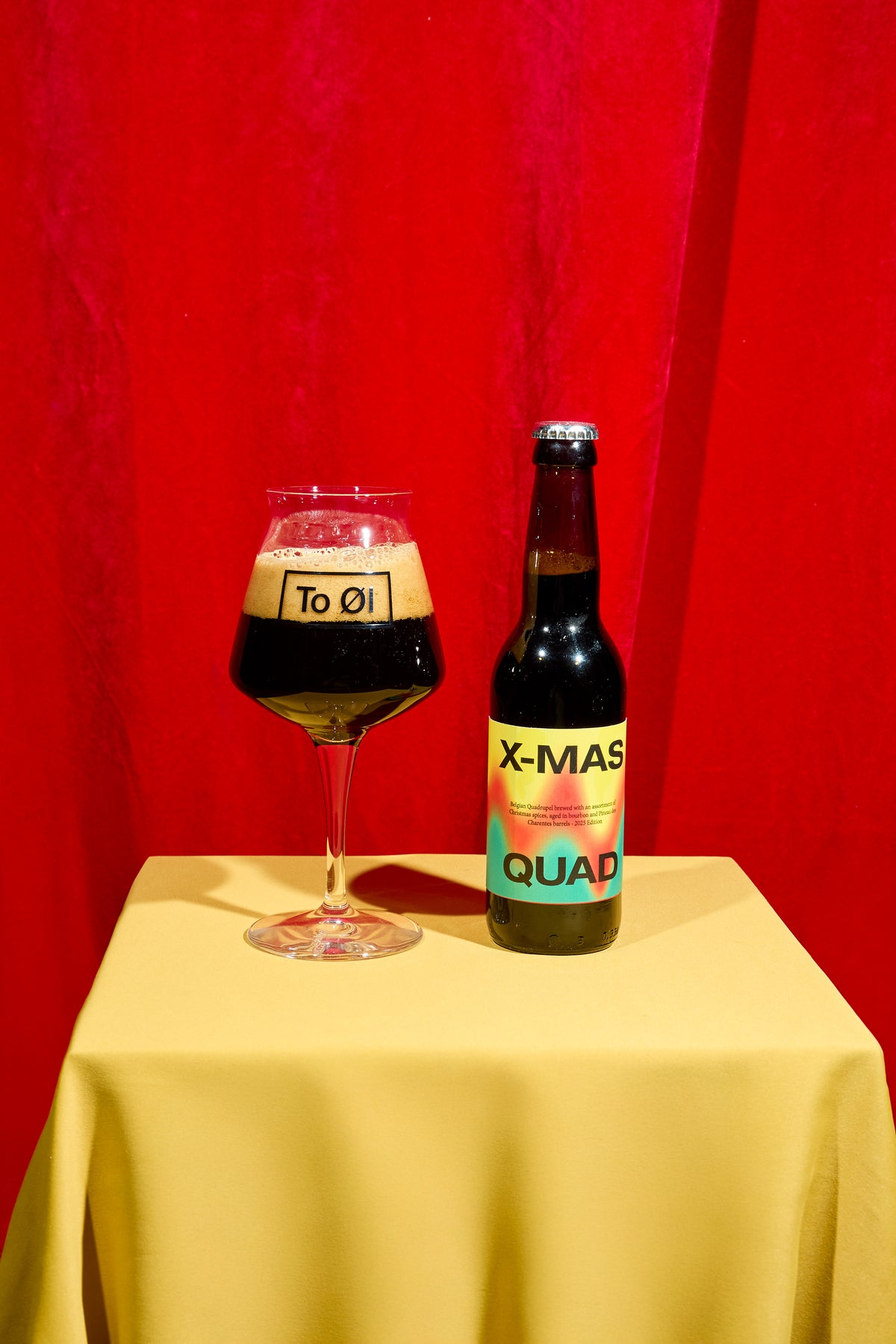 X-Mas Quad 2025 beer bottle and poured beer in a glass on a yellow table cloth and red curtain in the background.