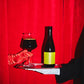 A dark glass bottle of Yule M#@!*?k Bourbon BA by To Øl with a bright yellow label sits on a silver tray held by a hand in a white glove, next to a filled wine glass, against a red curtain backdrop.