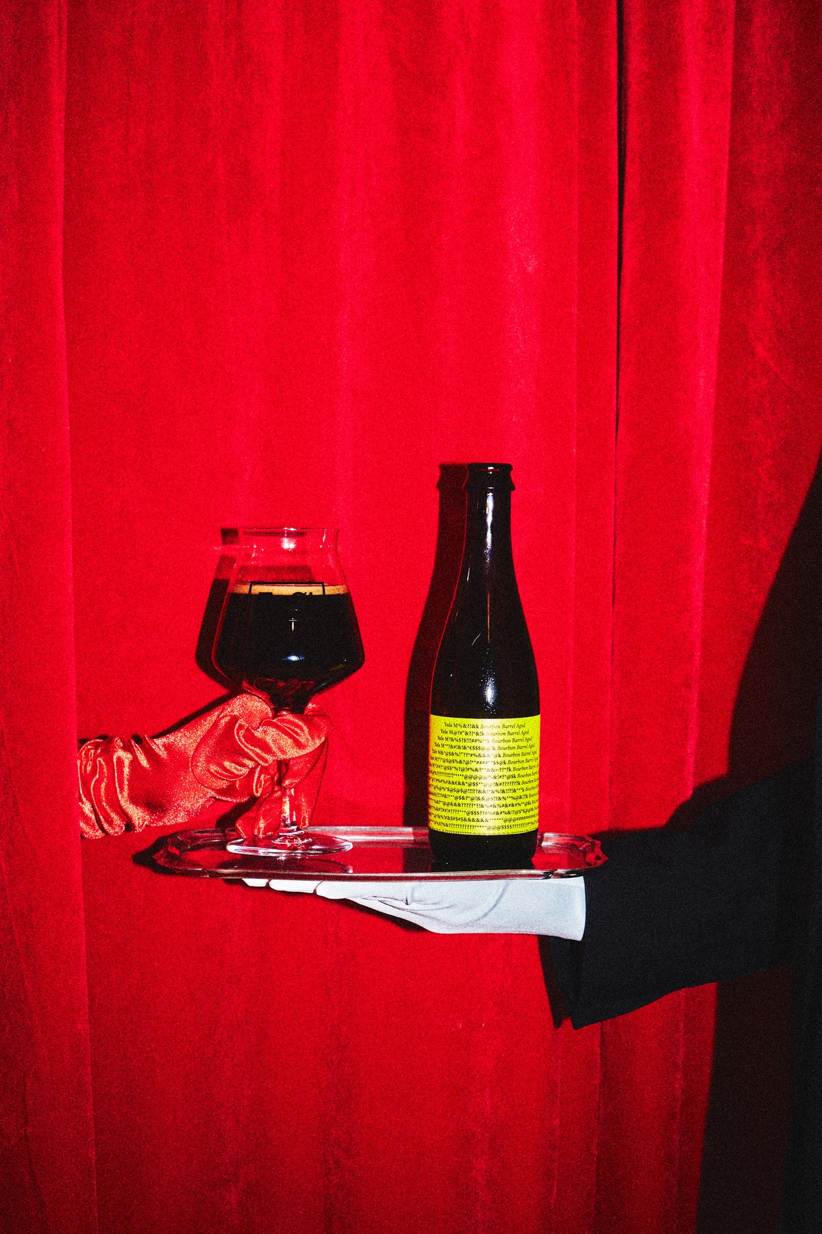 A dark glass bottle of Yule M#@!*?k Bourbon BA by To Øl with a bright yellow label sits on a silver tray held by a hand in a white glove, next to a filled wine glass, against a red curtain backdrop.