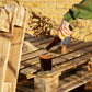 A person in a green sweatshirt pours a dark brown beverage from a Yule Malt BA - 2024 bottle into a clear glass resting on rustic wooden pallets, with a yellow brick wall in the background.