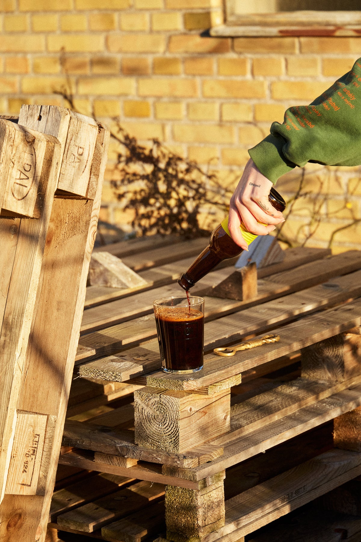 A person in a green sweatshirt pours a dark brown beverage from a Yule Malt BA - 2024 bottle into a clear glass resting on rustic wooden pallets, with a yellow brick wall in the background.