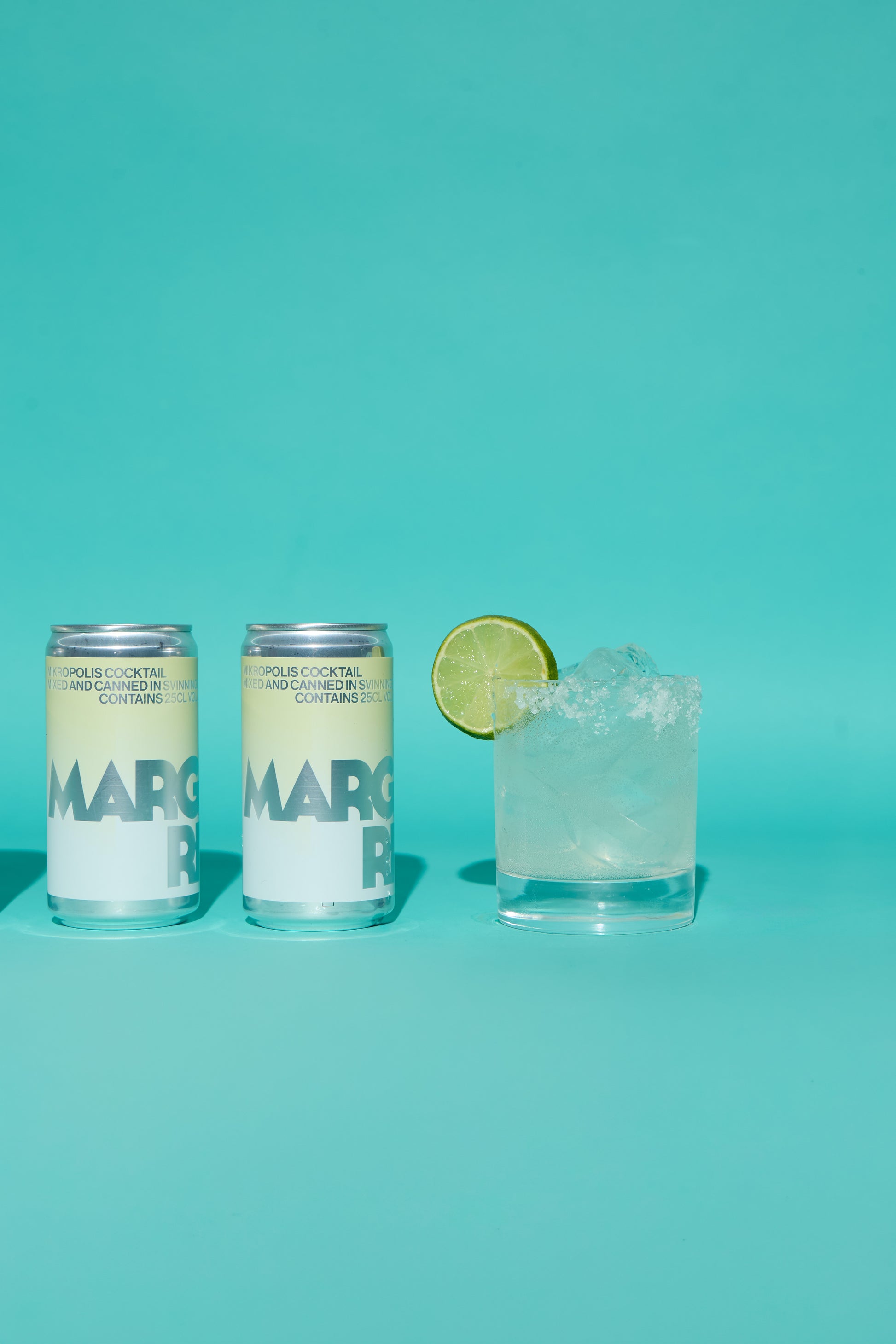 Two cans of Mikropolis Margaritas on a turquoise background with a glass of margarita and lime.