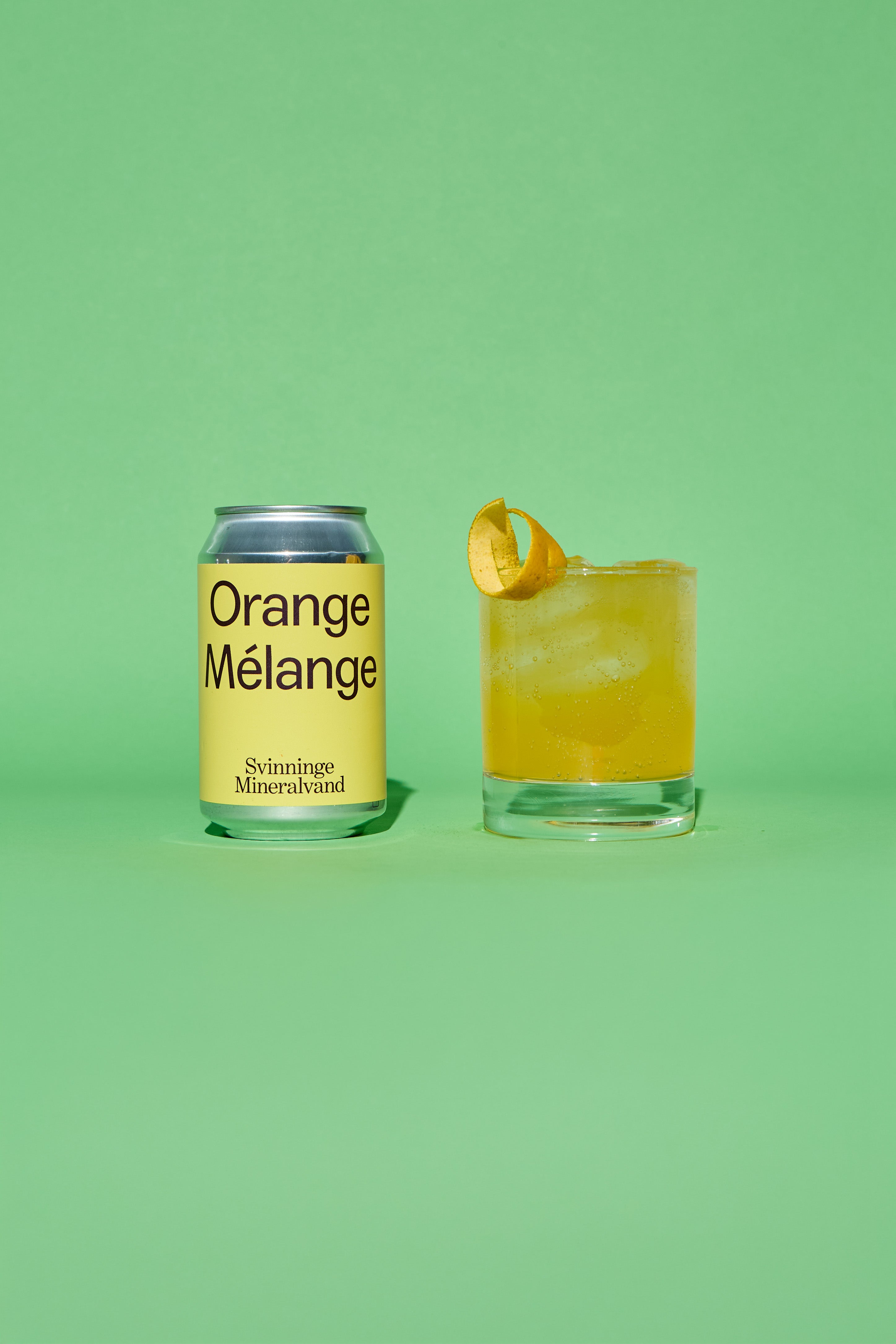 Yellow and silver can of 'Orange Melange' and a glass of orange cocktail on a green background.